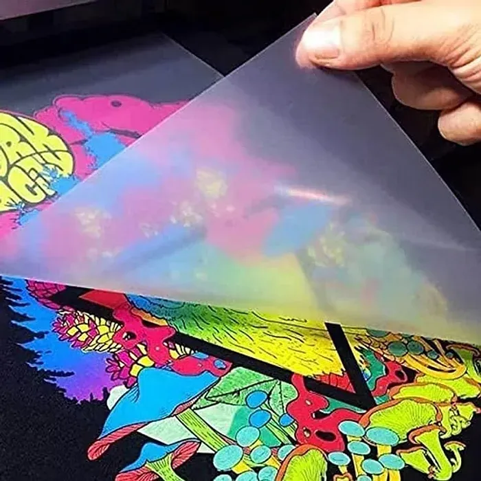 Filme transparente sendo puxado da camiseta preta, deixando a estampa colorida fixada no tecido